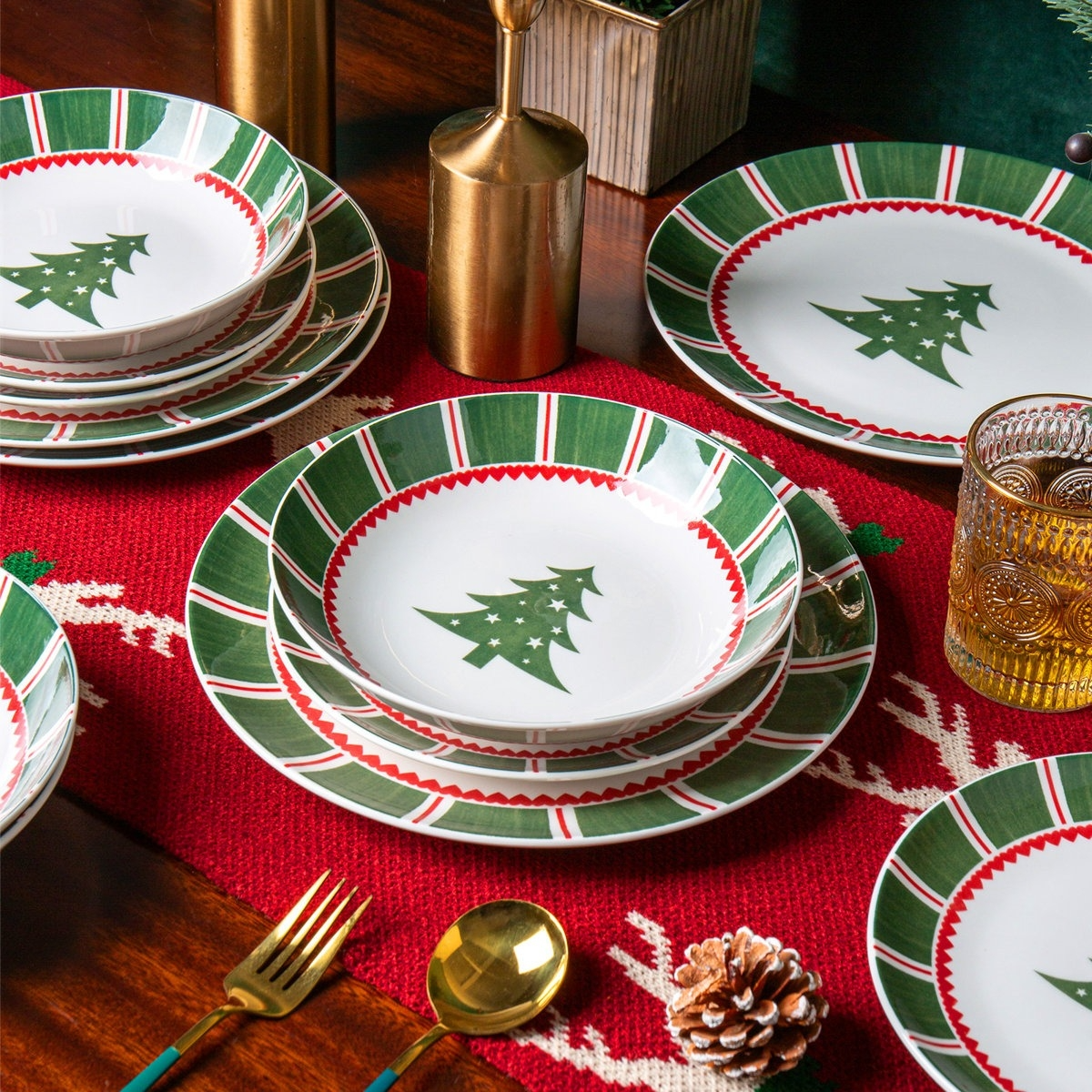 Christmas-themed tableware set with plates and bowls featuring green and red patterns and Christmas tree designs, accompanied by gold utensils and a textured glass on a festive red table runner.