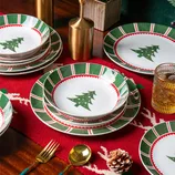 Christmas-themed tableware set with plates and bowls featuring green and red patterns and Christmas tree designs, accompanied by gold utensils and a textured glass on a festive red table runner.