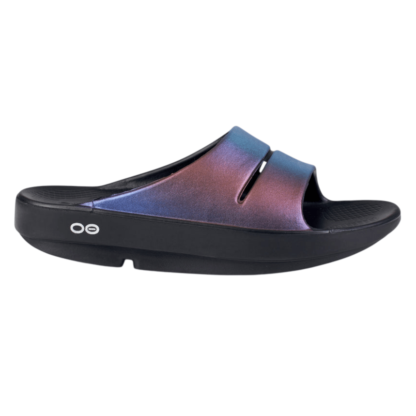 A black slide sandal with a shimmering, multicolored strap. The sandal features a thick sole with a simple branding logo on the side.