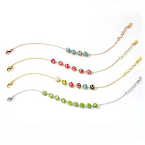 Four custom enamel slider charm bracelets with names \“EMMA,\“ \“JAYDEN,\“ and \“GABRIELLA\“ displayed in colorful circular charms on chains of different colors and clasp styles.