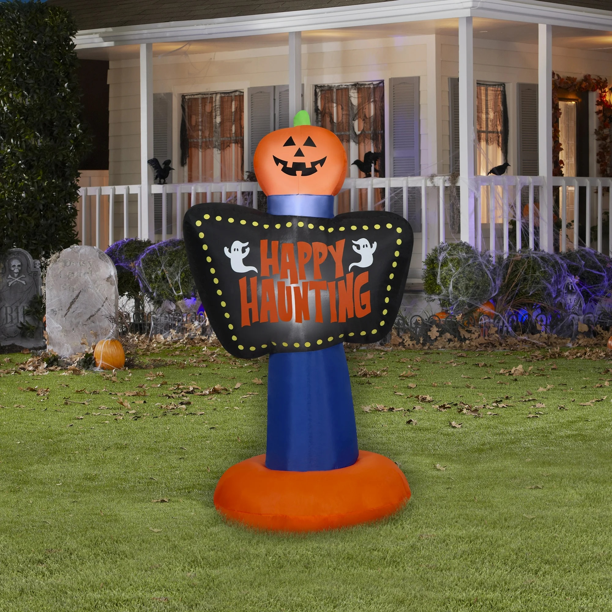 An inflatable Halloween decoration with a pumpkin head, a sign that reads \“Happy Haunting,\“ and a ghost motif, set on a lawn with other spooky decor in the background.
