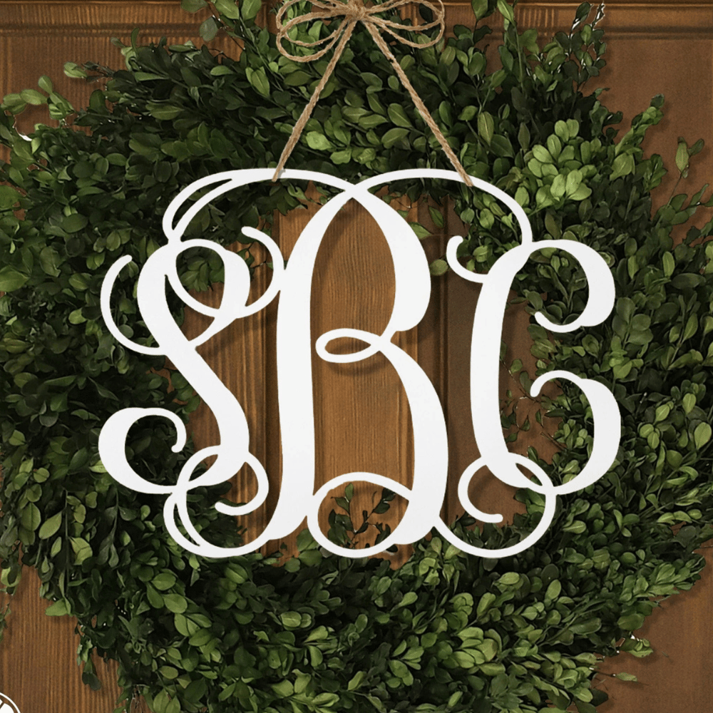 A green wreath with a decorative monogram \“SBC\“ in white, elegantly hung with a twine bow against a wooden background.