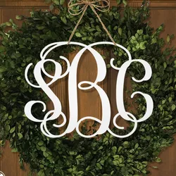 A green wreath with a decorative monogram \“SBC\“ in white, elegantly hung with a twine bow against a wooden background.