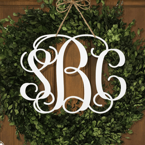 A green wreath with a decorative monogram \“SBC\“ in white, elegantly hung with a twine bow against a wooden background.