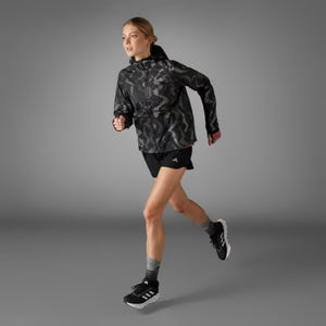 A woman is jogging in a black patterned jacket, black shorts, black sneakers with white stripes, and gray socks.