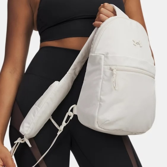 A person in athletic wear is holding a white sling backpack with a front zipper pocket. The outfit includes a black sports bra and black shorts with mesh panels.