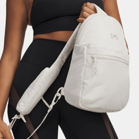 A person in athletic wear is holding a white sling backpack with a front zipper pocket. The outfit includes a black sports bra and black shorts with mesh panels.