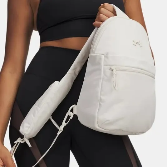 A person in athletic wear is holding a white sling backpack with a front zipper pocket. The outfit includes a black sports bra and black shorts with mesh panels.