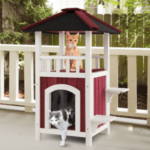 A two-level wooden cat house with a red and white color scheme features a cat sitting on the top level and another emerging from the bottom entrance.
