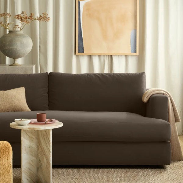 A dark brown sofa is paired with a light-colored throw and cushion. A round marble side table holds a cup and bowl. A decorative vase and abstract framed artwork hang in the background, complementing the neutral tones.