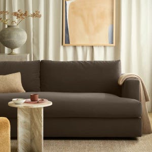 A dark brown sofa is paired with a light-colored throw and cushion. A round marble side table holds a cup and bowl. A decorative vase and abstract framed artwork hang in the background, complementing the neutral tones.
