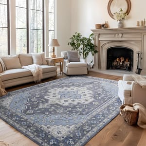 A living room features a blue and white patterned area rug, surrounded by a beige sofa, armchairs, a wooden side table, a potted plant, and a fireplace adorned with a mirror and decorative items.