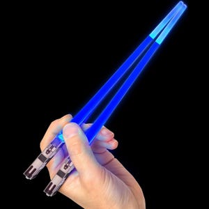 Blue light saber chopsticks are held in a hand, featuring handles designed to resemble the hilts of lightsabers. They emit a bright blue glow along the length of each chopstick.