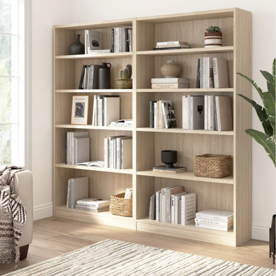 Light wood bookshelves with various books, decorative vases, plants, and woven baskets. A neutral-toned rug and a cozy blanket on a nearby sofa enhance the space.