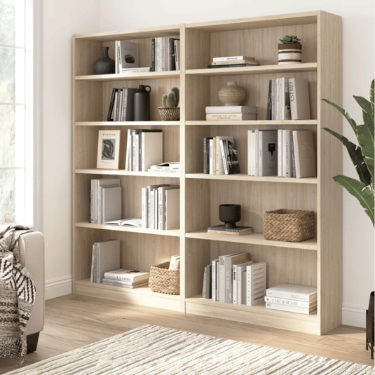 Light wood bookshelves with various books, decorative vases, plants, and woven baskets. A neutral-toned rug and a cozy blanket on a nearby sofa enhance the space.