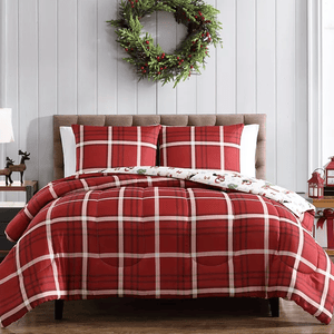 A bed with red and white plaid bedding, two pillows, and a reversible comforter featuring a festive design. A green wreath hangs on a light gray wall above, and decorative items, like a reindeer and lanterns, are on the side table.