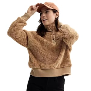 A woman is wearing a tan, plush, quarter-zip pullover with long sleeves and a ribbed hem, paired with a matching peach baseball cap.