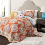 The 5-piece reversible queen quilt set features an orange and blue paisley design, includes a quilt, two shams, and two decorative pillows, set on a bed with a beige headboard and accompanied by a blue lamp on a wooden nightstand.