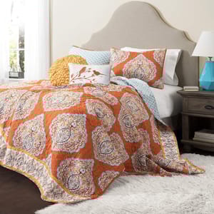 The 5-piece reversible queen quilt set features an orange and blue paisley design, includes a quilt, two shams, and two decorative pillows, set on a bed with a beige headboard and accompanied by a blue lamp on a wooden nightstand.