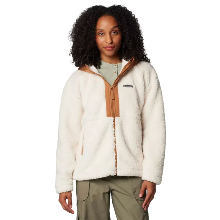A woman models a Columbia Hooded Sherpa Jacket, featuring a fluffy, white sherpa fleece with a camel-colored zipper and collar accents, and functional side pockets. The model combines the jacket with khaki pants and a light green undershirt.