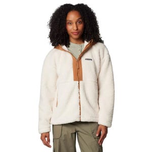 A woman models a Columbia Hooded Sherpa Jacket, featuring a fluffy, white sherpa fleece with a camel-colored zipper and collar accents, and functional side pockets. The model combines the jacket with khaki pants and a light green undershirt.