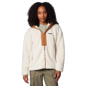 A woman models a Columbia Hooded Sherpa Jacket, featuring a fluffy, white sherpa fleece with a camel-colored zipper and collar accents, and functional side pockets. The model combines the jacket with khaki pants and a light green undershirt.