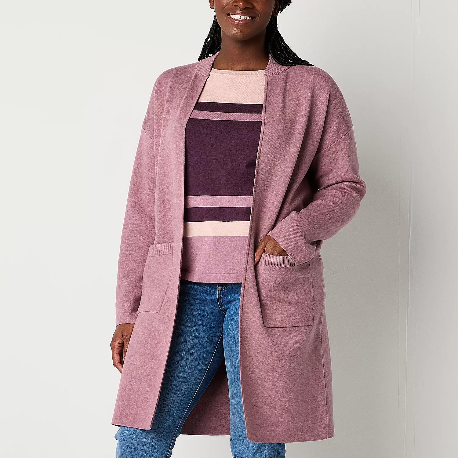 A woman is wearing a long, pink Liz Claiborne cardigan with front pockets, paired with a striped sweater and blue jeans.