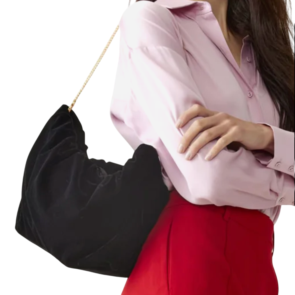 A woman in a pink blouse and red pants carries a soft, black velvet handbag with a gold chain strap.