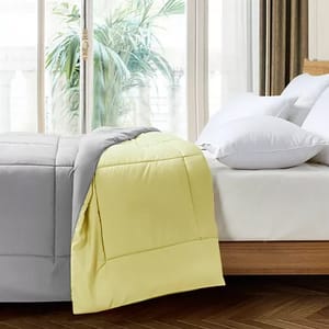 Reversible Down-Alt Queen Comforter with a gray side and a yellow side, shown on a bed beside white pillows, with a window and plants in the background.