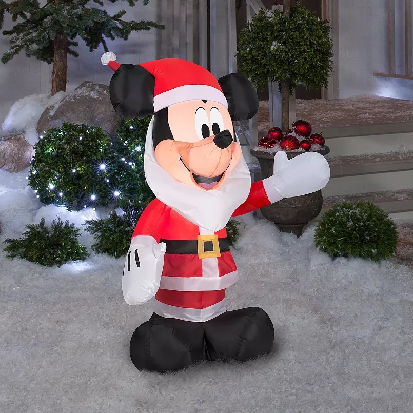 A Disney Mickey Mouse Santa inflatable dressed in a Santa suit with a white beard, red hat, and black belt, stands amidst snow and greenery with decorative lights in the background.