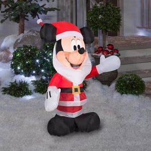 A Disney Mickey Mouse Santa inflatable dressed in a Santa suit with a white beard, red hat, and black belt, stands amidst snow and greenery with decorative lights in the background.