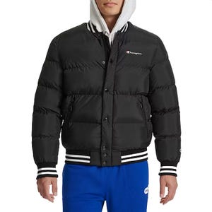 A black Champion Quilted Bomber Puffer Jacket featuring a snap button closure, side zip pockets, and ribbed cuffs and hem with white stripes. The jacket displays the Champion logo on the chest and is layered over a hooded sweatshirt.