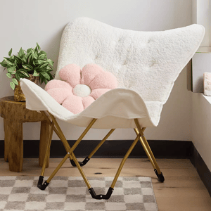 A fuzzy white chair with a pink flower-shaped cushion next to a plant on a wooden stool over a patterned rug.