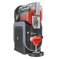 A Ninja SLUSHi maker is shown, designed to make frozen beverages, with a spout dispensing a red slush into a glass, garnished with a strawberry.