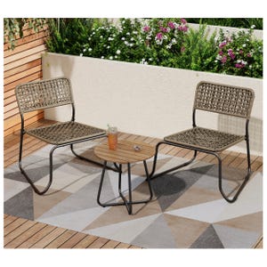 Outdoor seating set with two woven chairs and a small round wooden table on a geometric-patterned outdoor rug, surrounded by greenery and flowers.