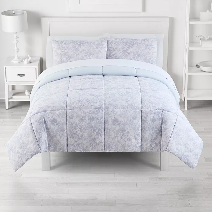 A light blue and white comforter set with a subtle floral pattern is displayed on a neatly made bed. The room features a white nightstand with a lamp and décor, and a shelving unit with various items, all against a minimalist white backdrop.