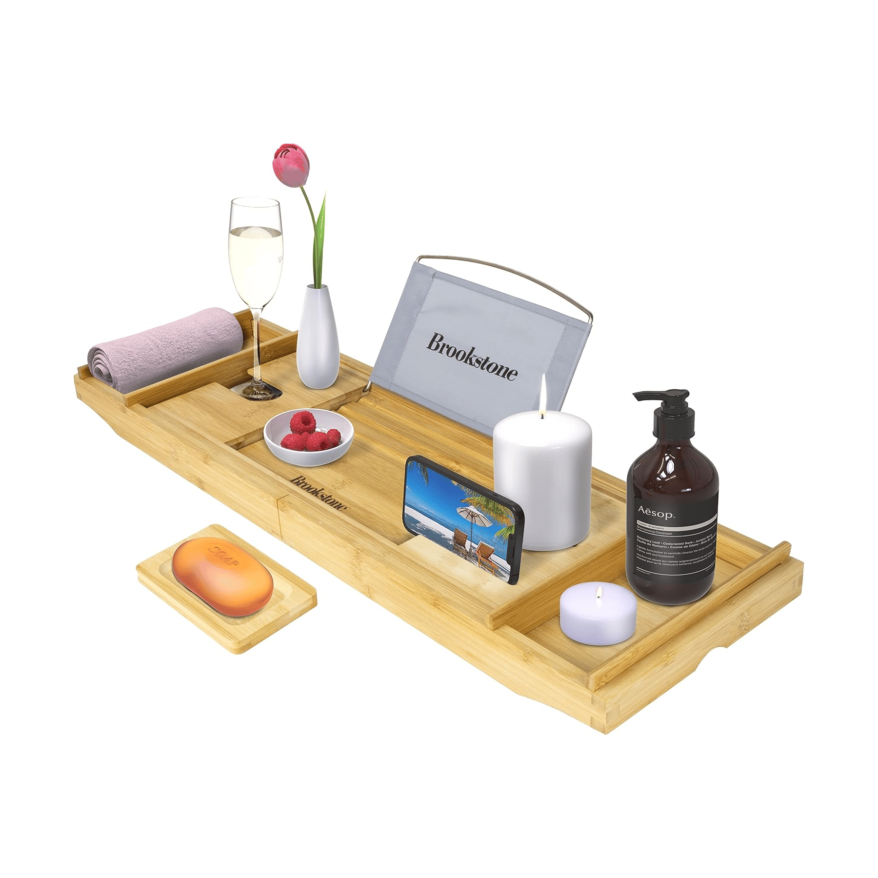 Bamboo bathtub tray with compartments holding a towel, wine glass, vase with a flower, bowl of raspberries, tablet stand, phone, candle, Aesop bottle, and a soap dish.