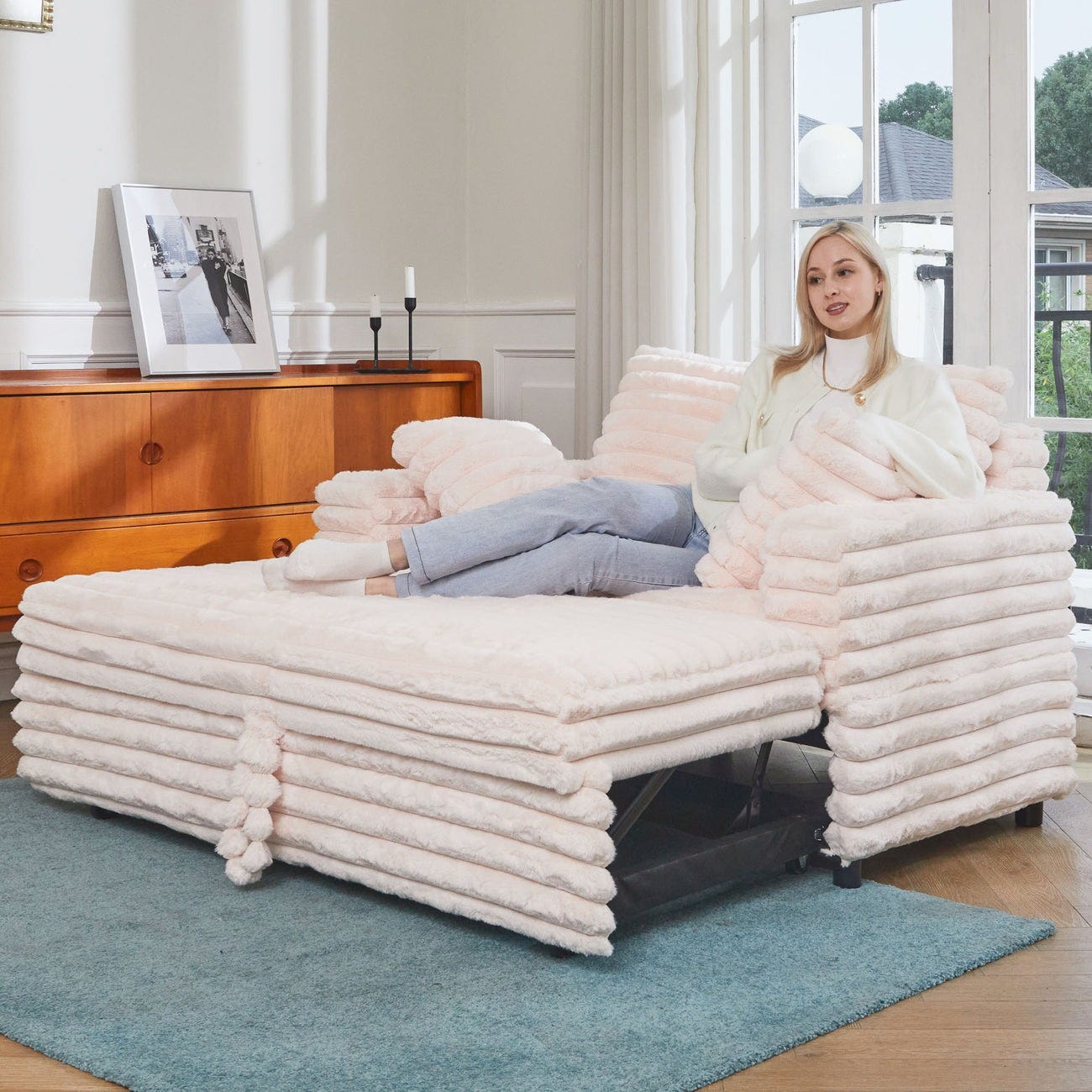 A woman sits on a plush, pale pink, stackable lounge mattress with matching cushions on a light blue rug, featuring a versatile design that combines seating and a fold-out bed.