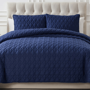 A navy blue 3-piece king quilt set featuring a quilted design with two matching pillow shams. The headboard is tufted and upholstered in a light, neutral color.