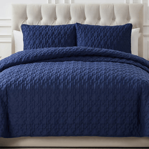 A navy blue 3-piece king quilt set featuring a quilted design with two matching pillow shams. The headboard is tufted and upholstered in a light, neutral color.