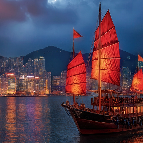 A traditional Chinese junk boat with red sails is illuminated against the backdrop of a city skyline at night, reflecting off the water.
