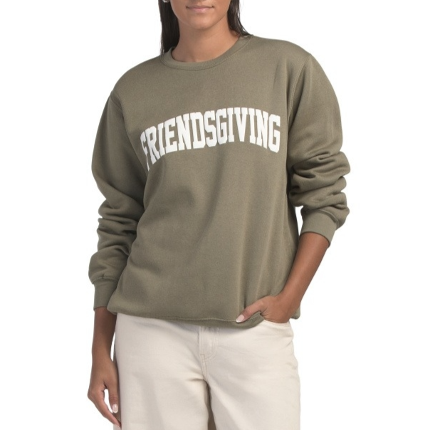 A person is wearing an olive green sweatshirt with \“FRIENDSGIVING\“ written in white on the front, paired with light beige pants.