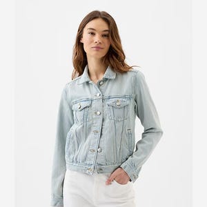 A light-wash denim jacket with a classic collar, button closures, flap pockets on the chest, and long sleeves. The jacket is styled on a woman paired with white pants.