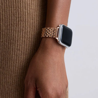 A person is wearing a smartwatch with a rose gold metal link bracelet on their wrist, against a background of brown fabric.