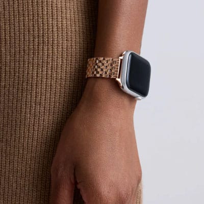 A person is wearing a smartwatch with a rose gold metal link bracelet on their wrist, against a background of brown fabric.