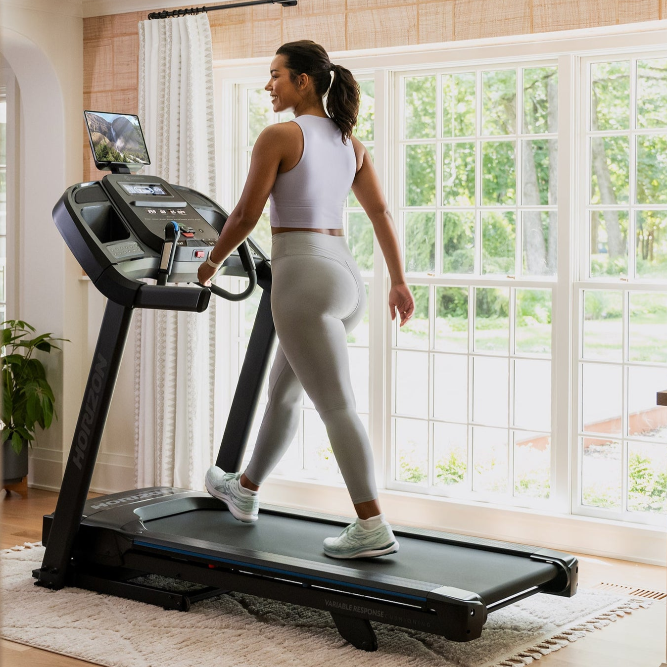 A person exercises on a Horizon treadmill positioned in a well-lit room with large windows.