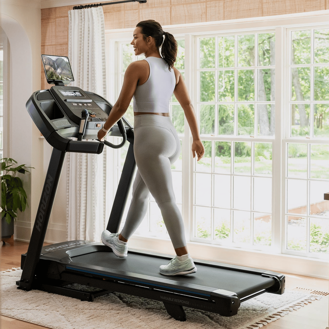 A person exercises on a Horizon treadmill positioned in a well-lit room with large windows.