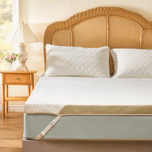 A bed features a memory foam topper with quilted cover and straps, alongside white quilted pillows and a wicker headboard, next to a wooden nightstand with a lamp and floral arrangement.