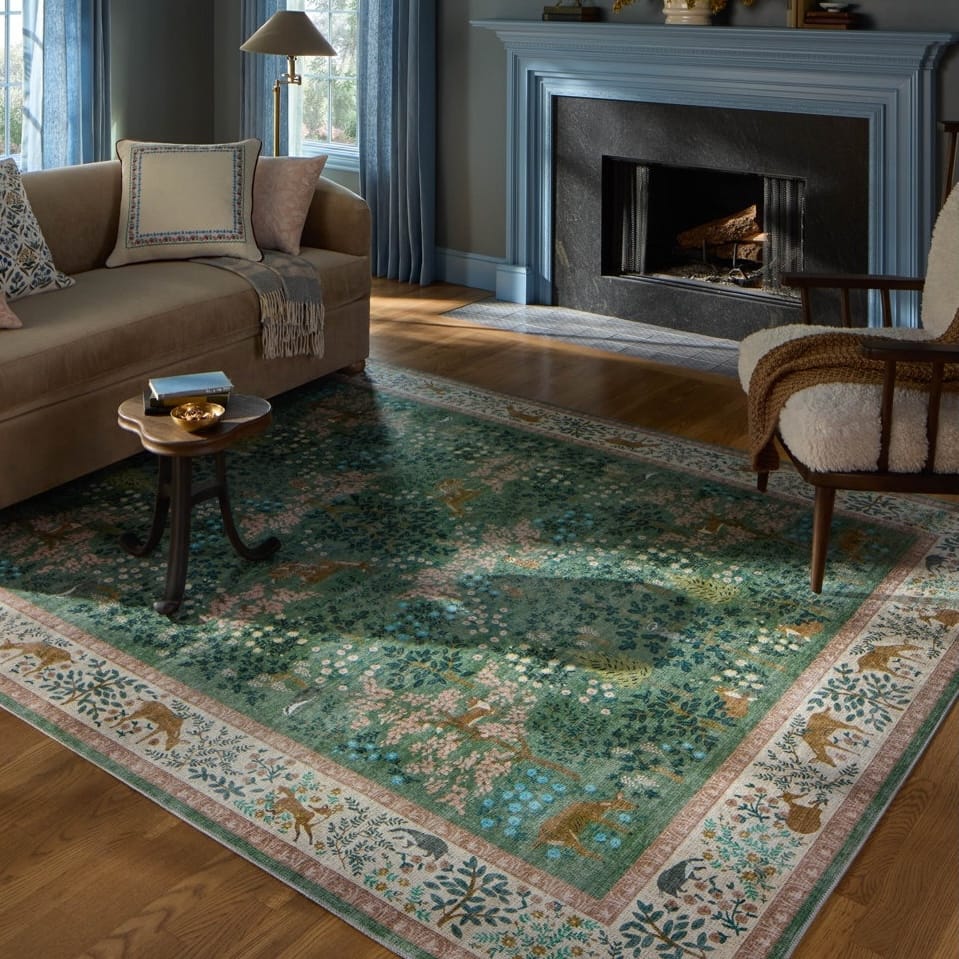 A living room with a beige sofa, small side table, and a vibrant green decorative rug featuring floral and animal motifs, placed on wooden flooring near a fireplace.