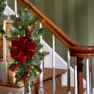 A faux-pine swag with a red bow, berries, pine cones, and lights is draped over a banister.